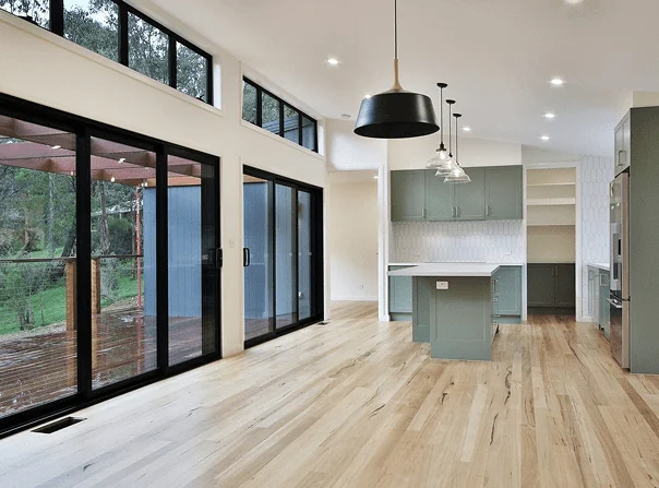 Modern open-plan kitchen and dining area by Coy’s Constructions, featuring timber flooring, custom sage green cabinetry, pendant lighting, and full-height sliding glass doors overlooking a timber deck and native garden.