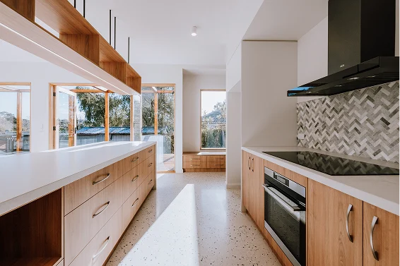 Contemporary kitchen interior with wood cabinetry, white countertops, herringbone tile backsplash, and built-in modern appliances – natural light from large windows enhances the space.